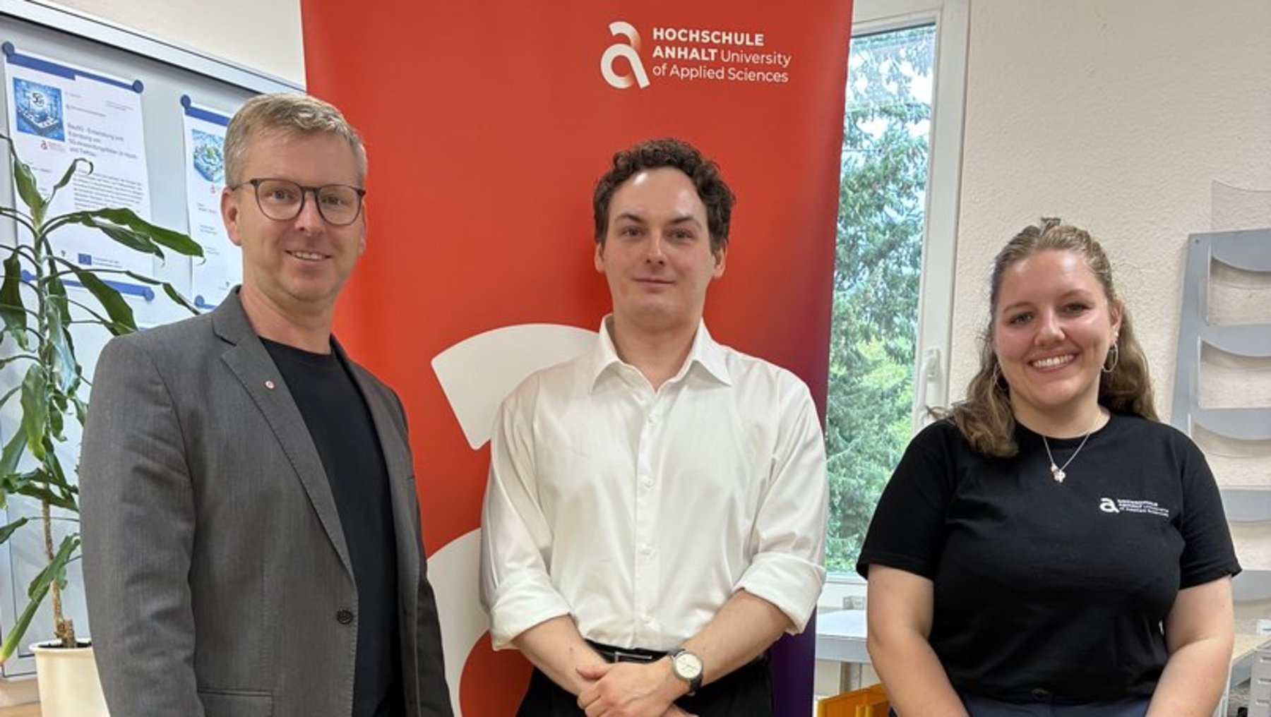 Drei Personen stehen lächelnd nebeneinander vor einem roten Banner mit dem Logo der Hochschule Aalen. Der Mann links trägt eine graue Jacke und Jeans, der Mann in der Mitte ein weißes Hemd, die Frau rechts ein dunkelblaues Shirt mit Logo und schwarze Hose.
