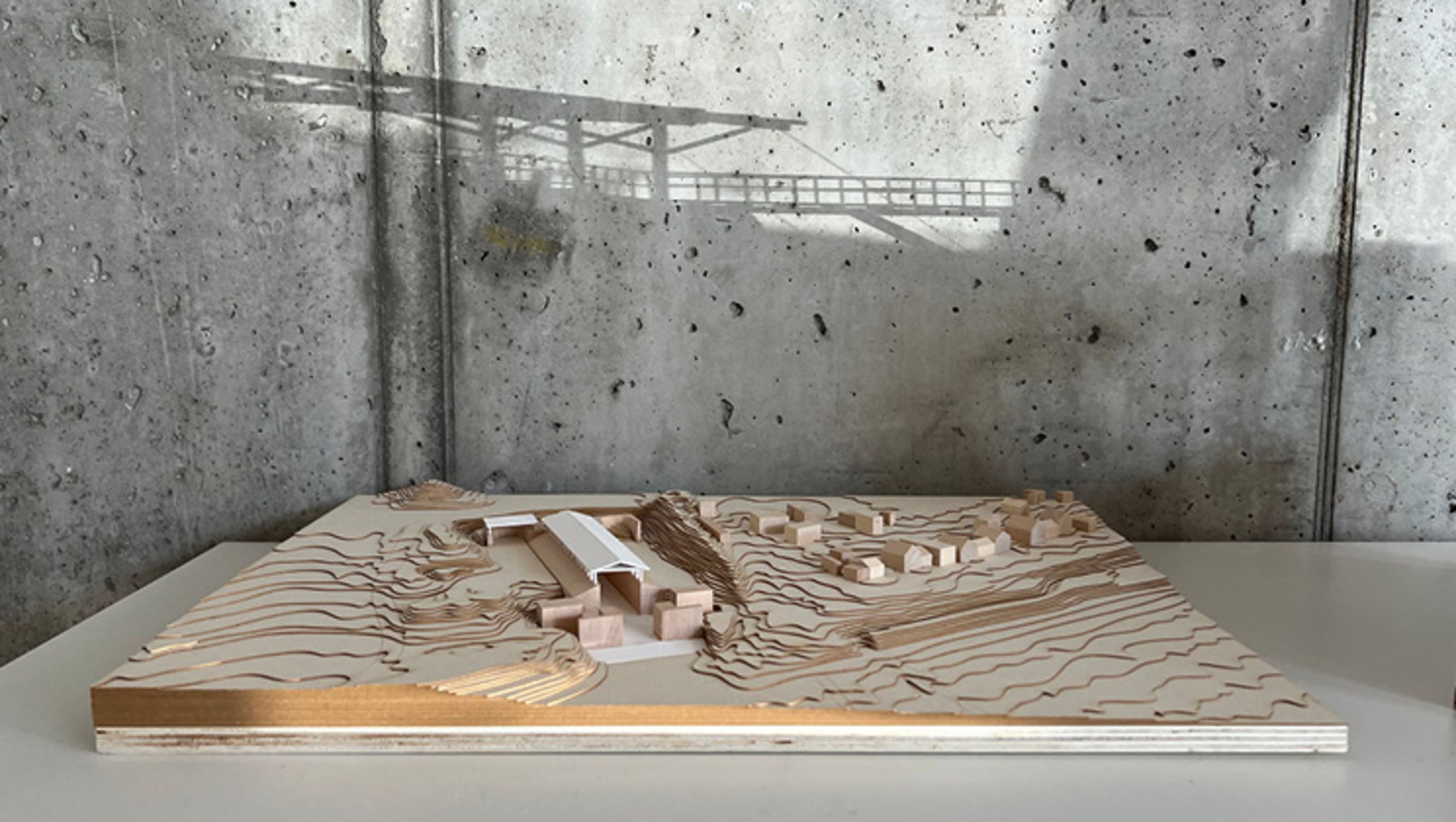 Holzreliefmodell eines Stadtteils mit Gebäuden, Straßen und wellenförmigem Gelände, das an einen Fluss oder eine Flusslandschaft erinnert. Im Hintergrund eine Brücke über dem Modell.