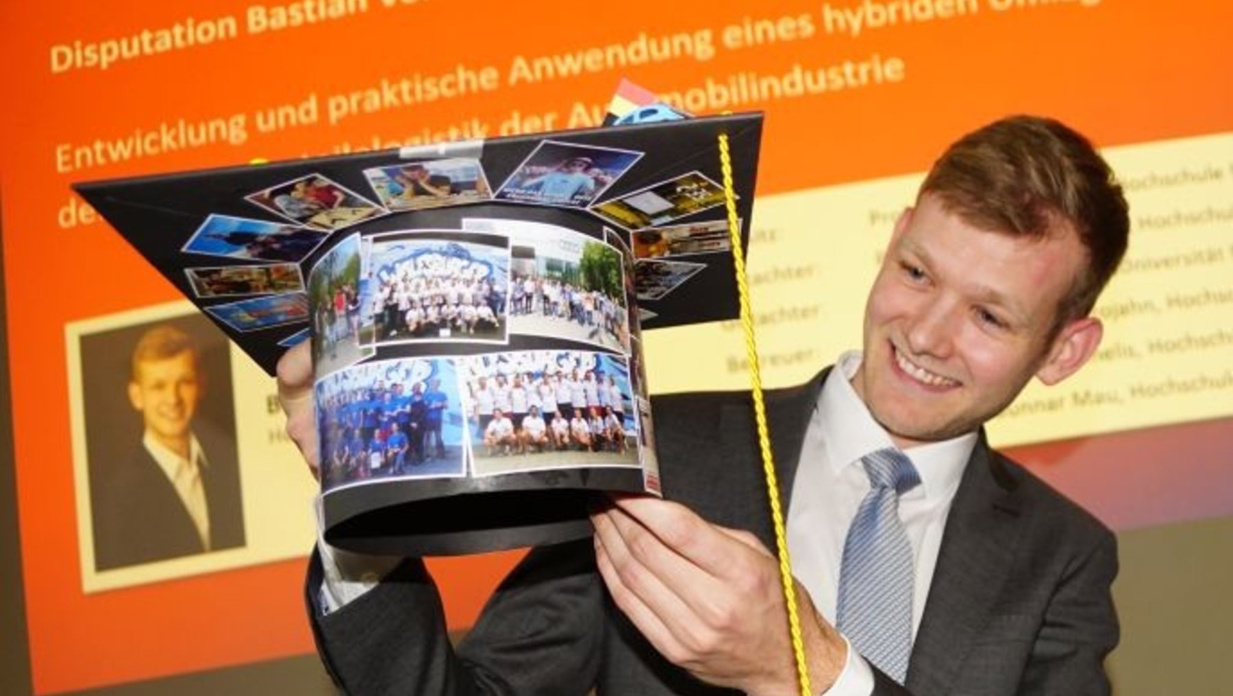 Ein lächelnder Mann hält eine Doktorhut-Kopie mit Fotos von Gruppen und einer Landschaft, die mit seiner Dissertation „Entwicklung und praktische Anwendung eines hybriden Umlagerungsverfahrens in der Automobilindustrie“ zusammenhängen.
