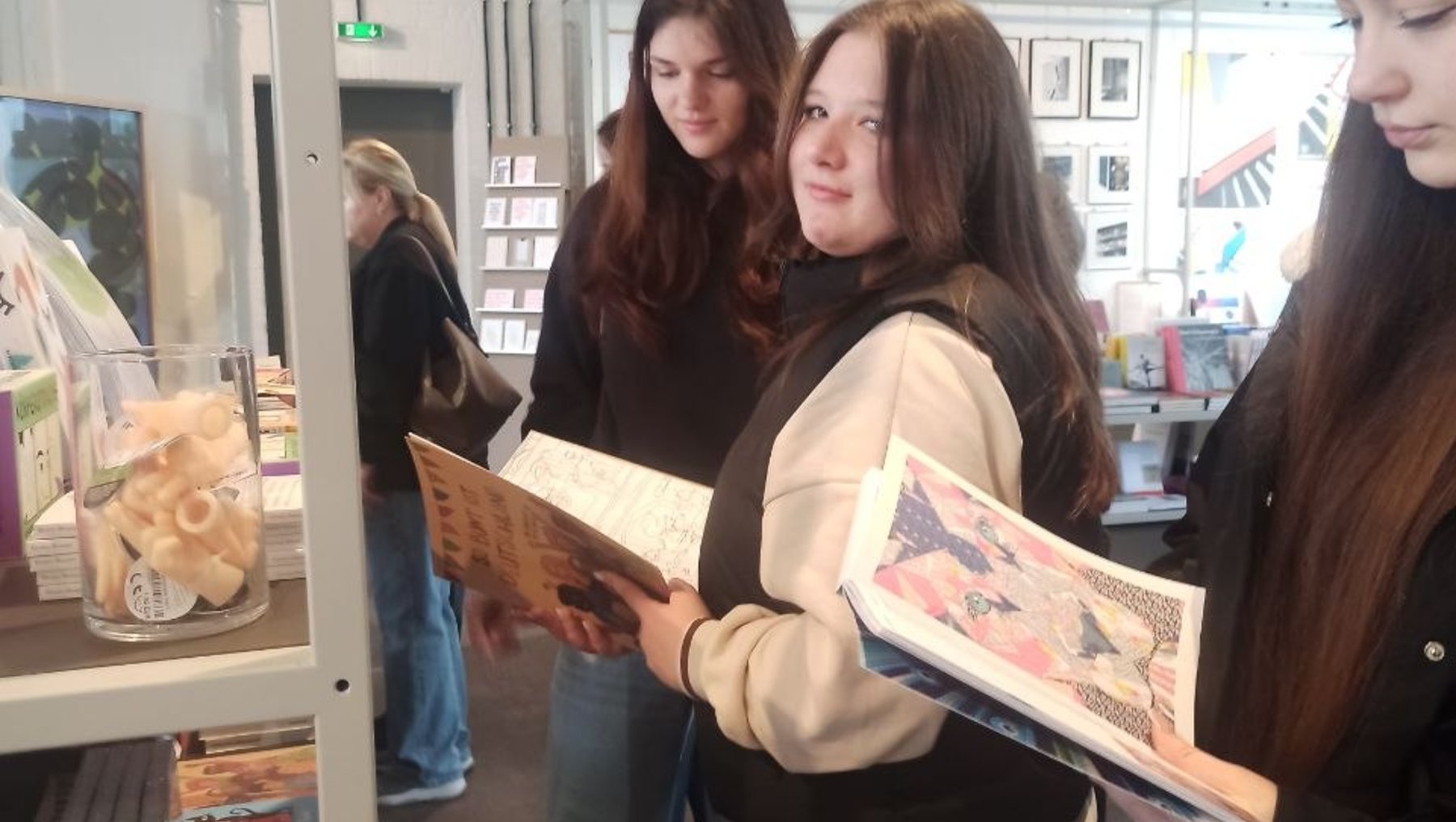 Drei Personen in einem Raum mit Regalen, die sich für Bücher interessieren: Eine Frau hält ein aufgeschlagenes Buch mit einer Illustration, zwei weitere schauen auf die Seite.