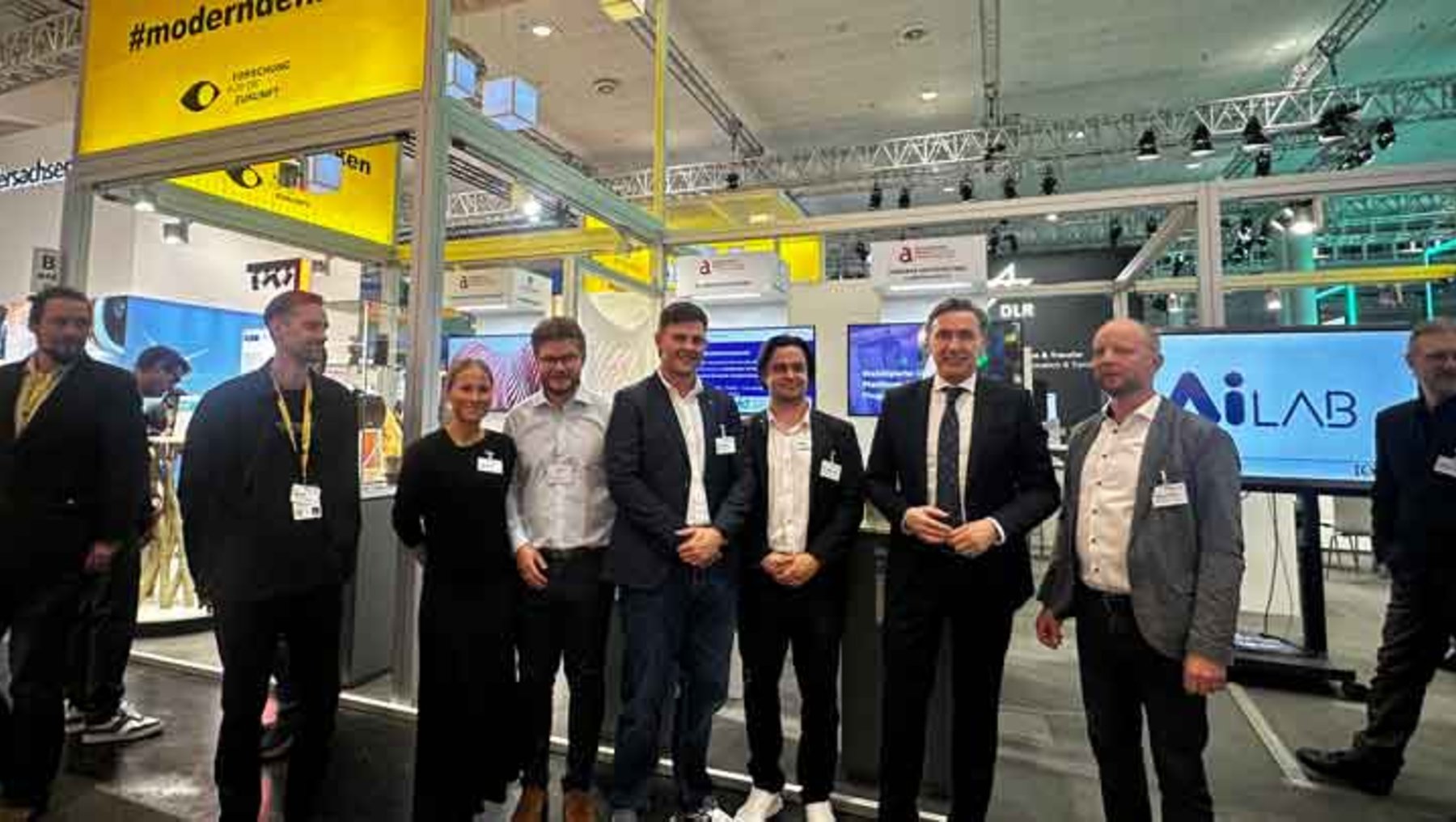 Gruppe von sieben Personen auf einer Messe, die lächelnd für ein Gruppenfoto posiert. Im Hintergrund sind Messestände mit Infoschildern wie „#moderninden“ und „AI LAB“ zu sehen.