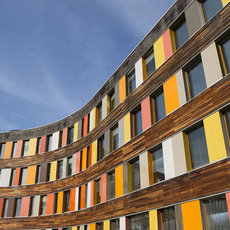 Moderne, kurvige Wohngebäude mit Holzfassade und farbigen Vertikalstreifen in Rot, Orange, Gelb und Weiß, die sich am Himmel abzeichnen.