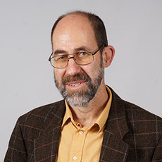 A man with glasses, a beard, and mustache, wearing a yellow shirt and a brown plaid blazer, smiling at the camera. The background is plain and light-colored.