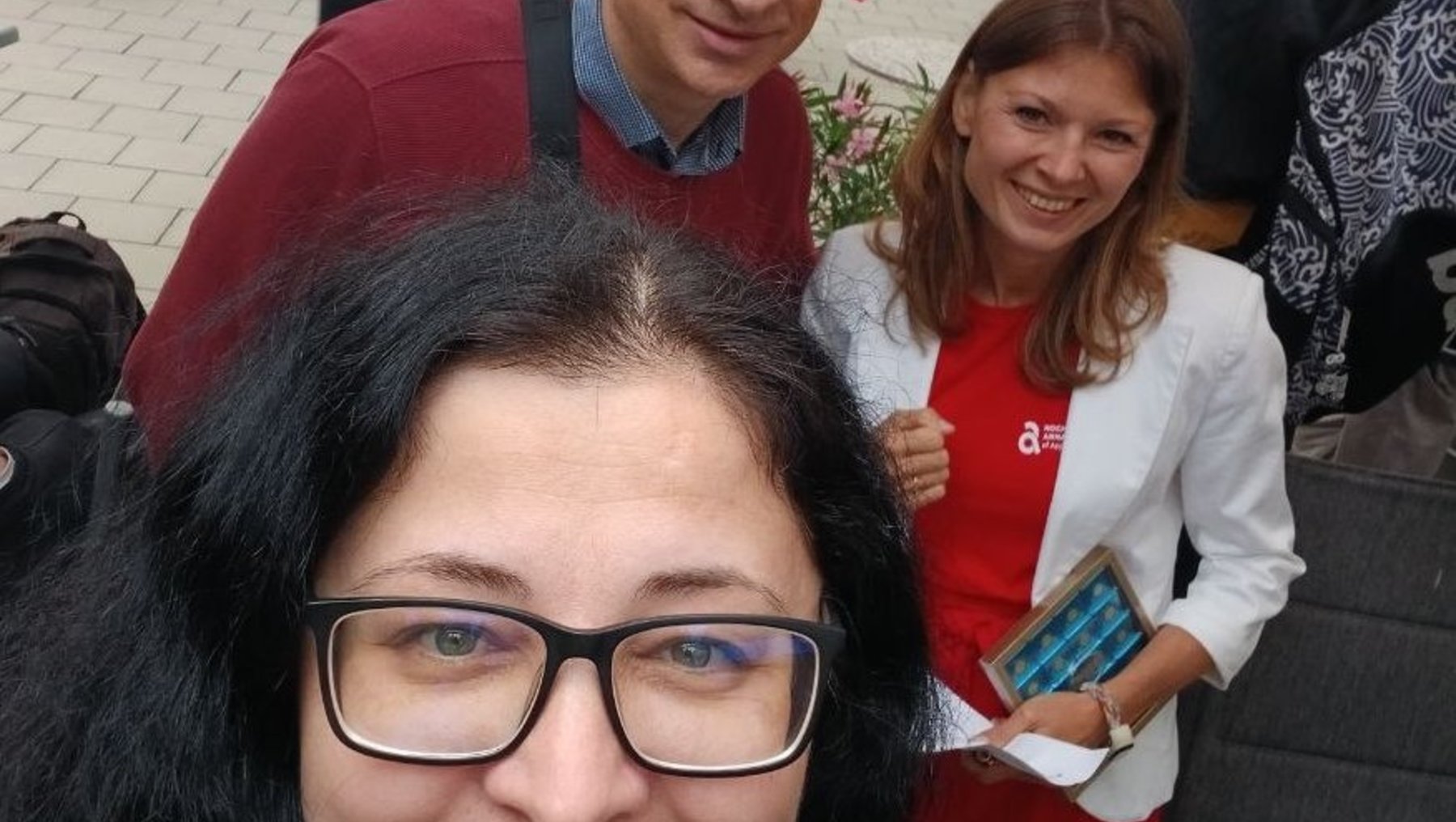 Drei Personen lächeln auf einem Selfie vor einem Pavillon mit Menschen im Hintergrund. Eine Frau mit Brille und dunklem Pulli im Vordergrund, ein Mann mit rotem Pulli und eine Frau mit weißem Oberteil und rotem Schal im Hintergrund.
