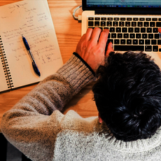 Person sitzt an einem Holzschreibtisch, arbeitet konzentriert an einem Laptop, daneben liegen ein Notizbuch mit Stift und eine Tablet.