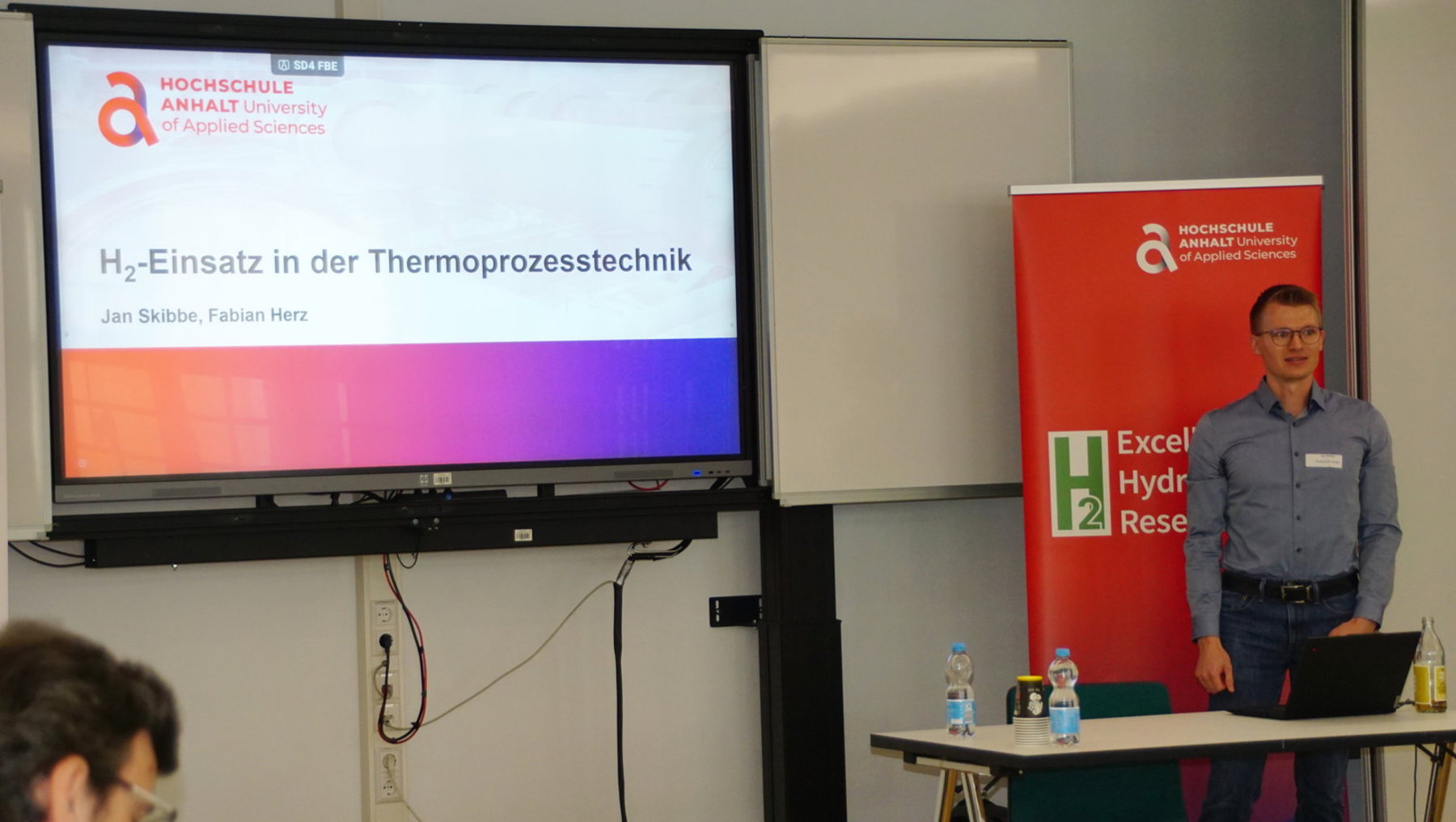 Vortragender steht vor einem Bildschirm mit Präsentation zum Thema Wasserstoff-Einsatz in der Thermoprozesstechnik. Zwei Referenten, Jan Skibbe und Fabian Herz, werden genannt. Ein Banner der Hochschule mit Logo und Schriftzug „Excellence in Hydrogen Research“ ist sichtbar.