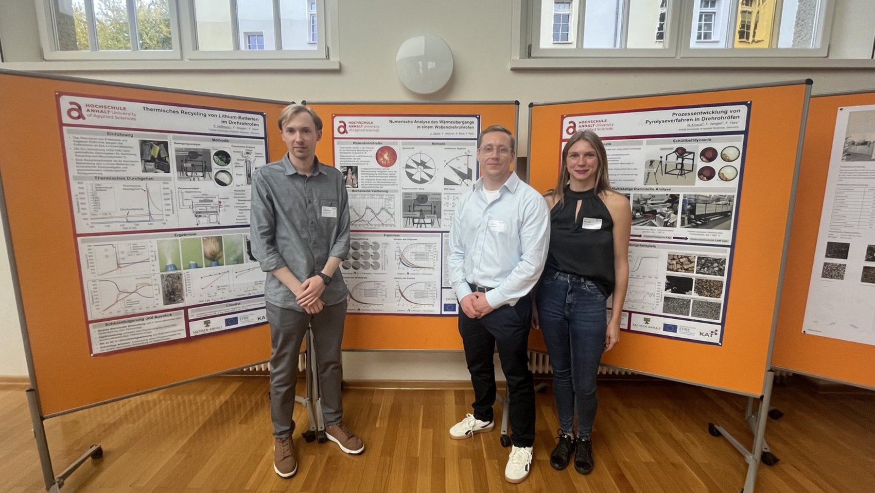 Drei Personen stehen vor drei orangefarbenen Posterwänden mit wissenschaftlichen Präsentationen zu astronomischen Themen. Jedes Poster zeigt Grafiken, Diagramme und Fotos von Himmelskörpern sowie Messdaten. Die Personen lächeln und tragen Namensschilder.