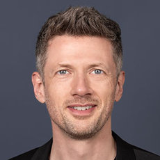 Lächelnder Mann mit kurz geschnittenen, hellen Haaren und blau-grauen Augen, der direkt in die Kamera blickt. Er trägt ein schwarzes Oberteil.