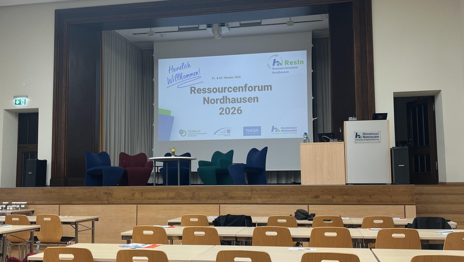 Leerer Seminarraum mit Tischen und Stühlen in U-Form, vorbereitet für das Ressourcenforum Nordhausen 2024. Vorne große Leinwand mit Veranstaltungstitel und Logo. Auf Tischen liegen Informationsmaterialien und Stifte.