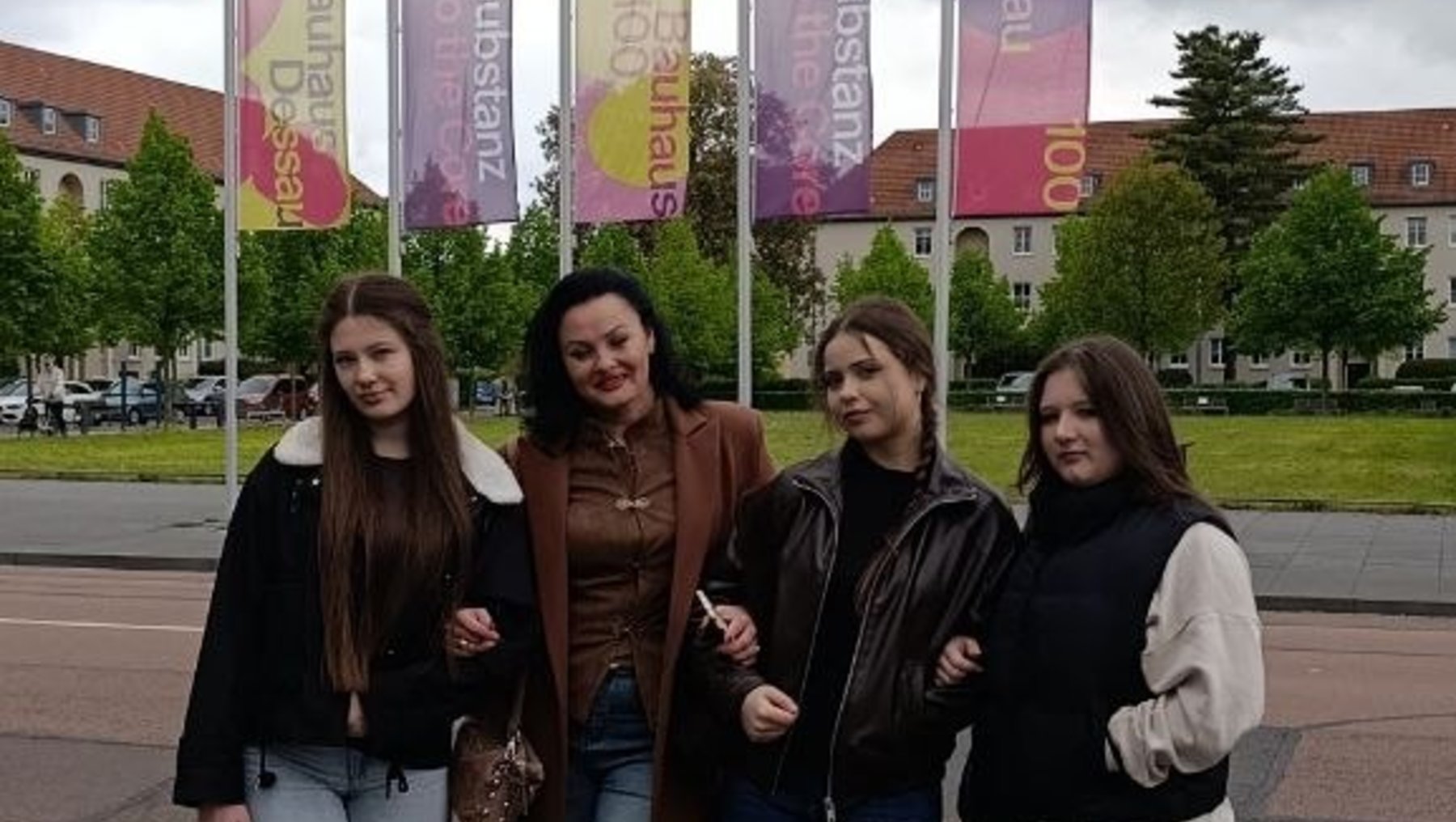 Vier Personen stehen lächelnd vor bunten Banner-Wänden mit dem Schriftzug „Messe“ oder ähnlichem Eventnamen, im Hintergrund ein Gebäude mit Rasenfläche und Bäumen.