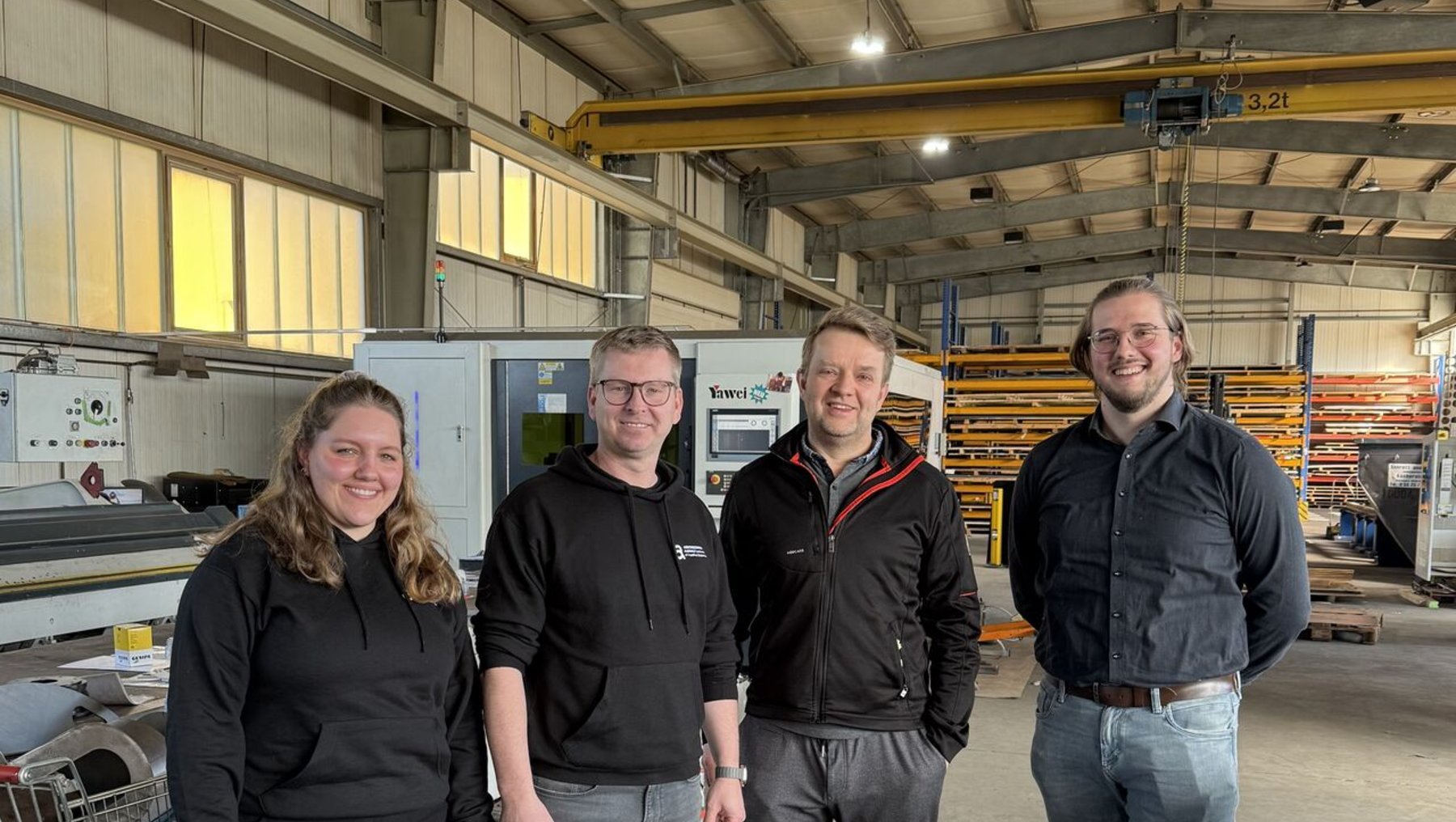 Vier Personen stehen lächelnd in einer Industriehalle mit Maschinen und Regalen im Hintergrund, darunter eine Frau mit langen Haaren und drei Männer in Arbeitskleidung, zwei davon mit Firmenlogos auf den Jacken.