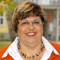 A smiling woman with short brown hair, wearing glasses, an orange top, and a colorful beaded necklace. She is standing outdoors with a building in the background.