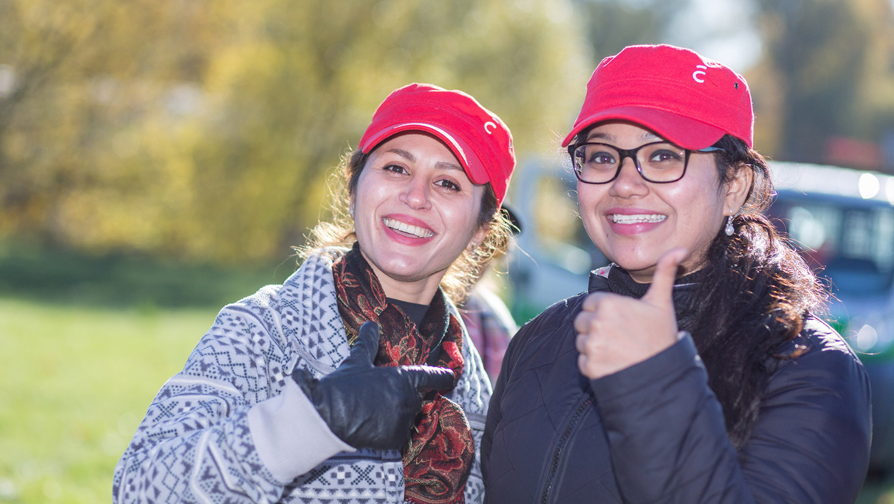 Zwei Frauen mit roten Baseballkappen lächeln in die Kamera. Sie tragen warme Kleidung und stehen im Freien, möglicherweise bei einem Outdoor-Event. Die eine Frau trägt eine gestreifte Jacke und einen Schal, die andere eine schwarze Jacke und eine Brille.