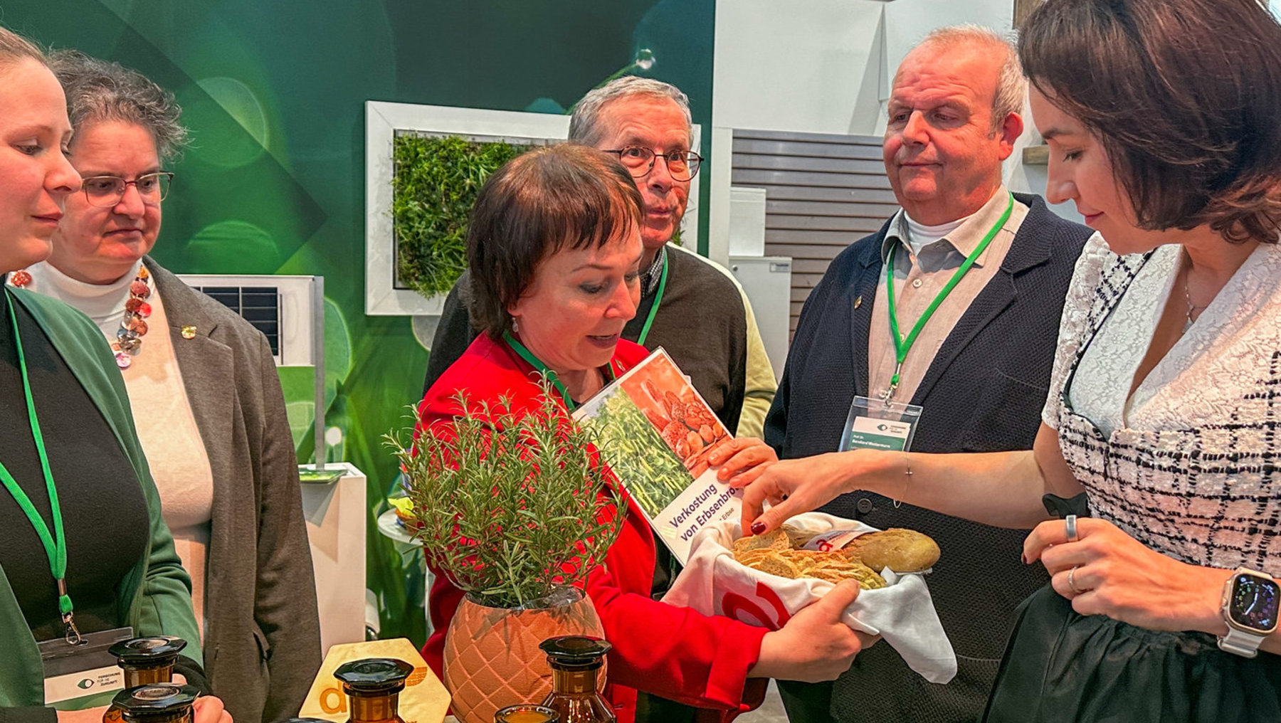 Eine Frau hält ein in Geschenkpapier eingewickeltes Paket mit einer Pflanze in einem roten Topf und einem Saatgutbeutel der Sorte „Erbsen“. Vier Personen stehen um sie herum und beobachten interessiert. Im Hintergrund sind Flaschen und ein Stand mit dem Namen „Wittenberg“ zu sehen.