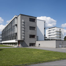 Moderne Bauhaus-Gebäude mit markantem Namen „Bauhaus“ an der Fassade, bestehend aus einem flachen, kubischen Hauptbau mit großen Fenstern und einem angrenzenden, hellen Anbau. Vor dem Gebäude liegt ein gepflegter Rasen.