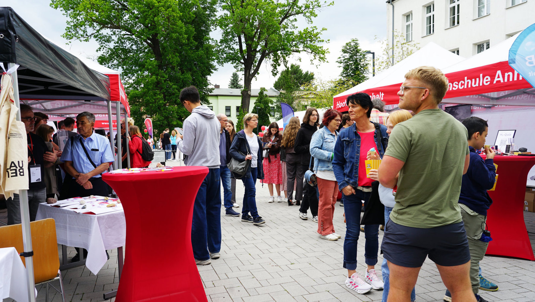 Menschen auf einer Außenveranstaltung mit mehreren Infoständen, darunter ein roter Tisch mit weißem Tischtuch und einem Schild „Ausbildung“. Besucher stehen in Gruppen, unterhalten sich oder informieren sich an den Ständen. Sonniges Wetter, Zelte und ein Gebäude im Hintergrund.