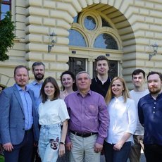 Eine Gruppe von Menschen steht vor einem historischen Gebäude mit einem großen, bogenförmigen Fenster. Die Gruppe besteht aus Männern und Frauen, die lächeln und für ein Gruppenfoto posieren. Sie sind vor einem grünen Rasen und Bäumen positioniert.