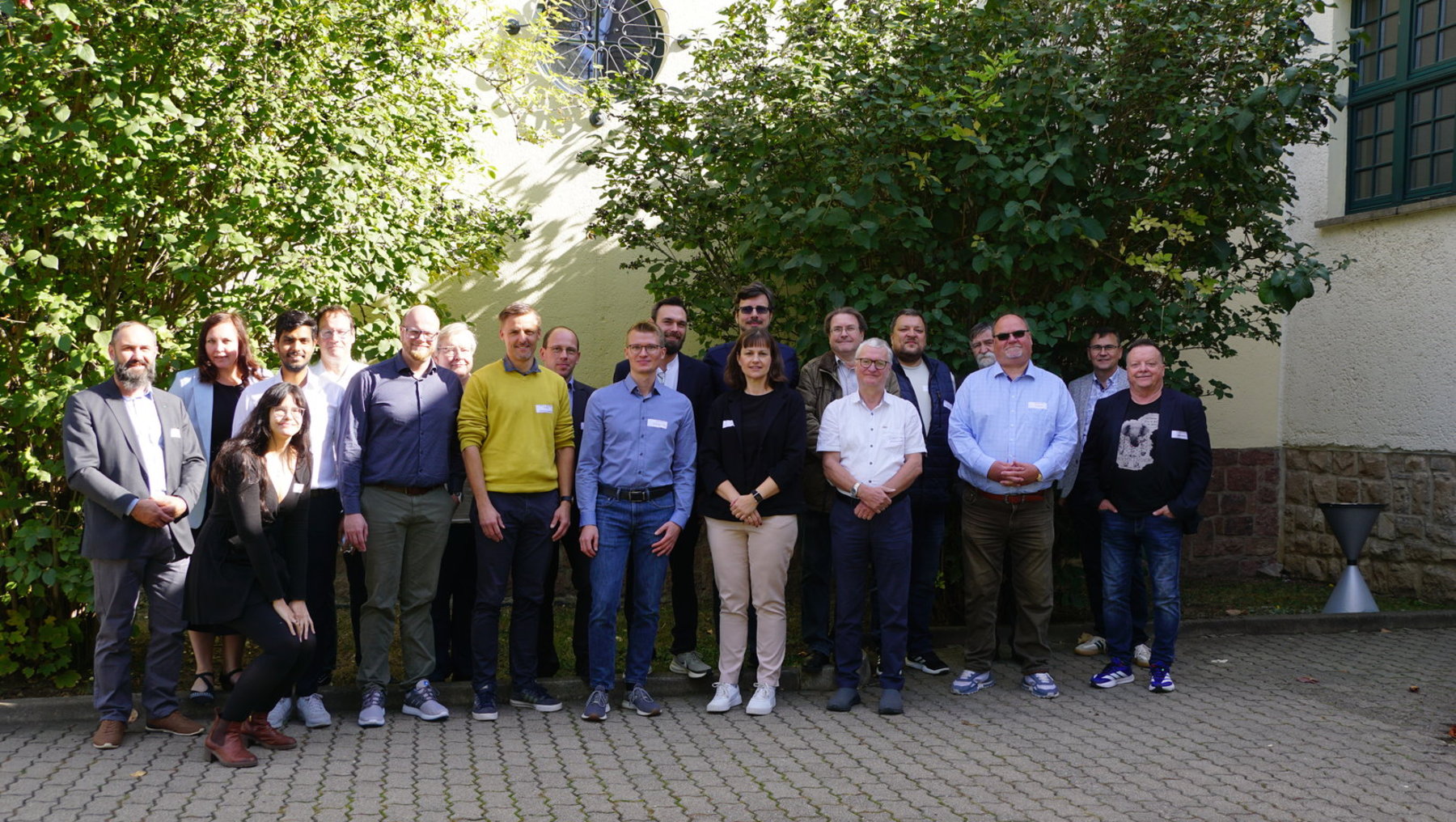 Gruppe von 18 Personen steht lächelnd im Freien für ein Gruppenfoto. Sie tragen verschiedene Kleidungsstücke wie Anzüge, Pullover und T-Shirts. Einige haben Namensschilder. Im Hintergrund sind Bäume und ein Gebäude mit Uhrturm sichtbar.