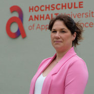 A woman in a pink blazer stands in front of a building with the sign "Hochschule Anhalt - University of Applied Sciences." She appears to be posing for the photo. The setting suggests an academic or professional environment.
