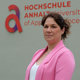 Eine Person mit hellrosa Blazer steht lächelnd vor einem grauen Hintergrund mit dem Logo und Schriftzug „Hochschule Anhalt – University of Applied Sciences“.