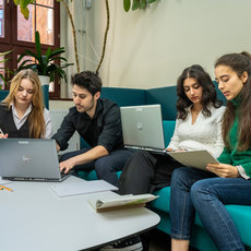 Four students are sitting on a blue couch in a study area. They are working on laptops and reviewing documents together. The setting appears to be a library or a study room with a table in front of them holding books and stationery.