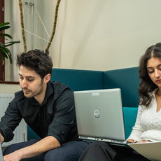 Vier Studenten sitzen auf einer blauen Couch und arbeiten gemeinsam an Laptops. Sie sind konzentriert und haben Notizbücher und Stifte auf einem Tisch vor sich liegen. Die Szene findet in einem hellen Raum mit Pflanzen im Hintergrund statt.