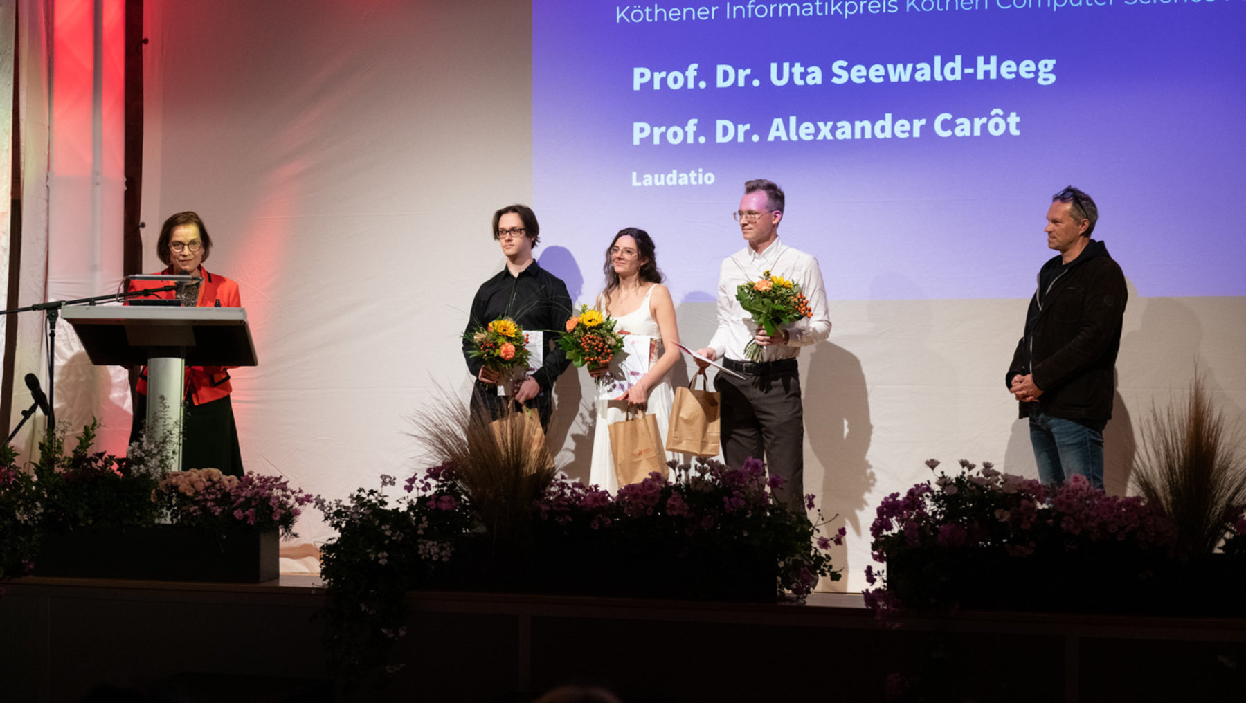 Preisträgerverleihung des Köthener Informatikpreises mit zwei Professoren im Fokus: Prof. Dr. Uta Seewald-Heeg und Prof. Dr. Alexander Carôt. Vier Personen auf Bühne, zwei davon halten Blumensträuße, eine hält eine Tüte. Laudatorin spricht am Rednerpult.