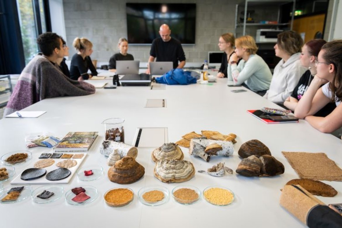 Ein Gruppenmeeting in einem modernen Raum. Auf dem Tisch stehen verschiedene Kuchen, Brote und Snacks. Die Teilnehmer sitzen um den Tisch herum und arbeiten an Laptops. Es herrscht eine entspannte Atmosphäre mit Essen und Diskussionen.