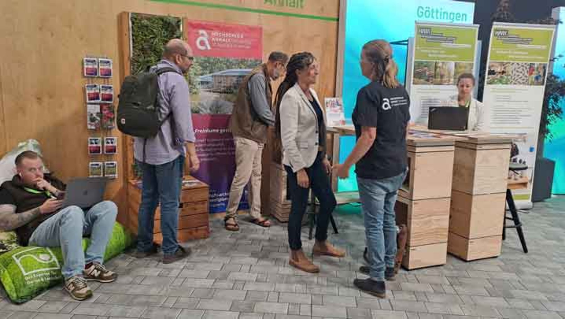 Drei Personen stehen vor einem Infostand mit Plakaten zu nachhaltiger Landwirtschaft und Biodiversität. Links sitzt eine Person auf dem Boden mit Rucksack. Der Stand zeigt das Logo der Universität Göttingen und thematisiert ökologische Projekte.