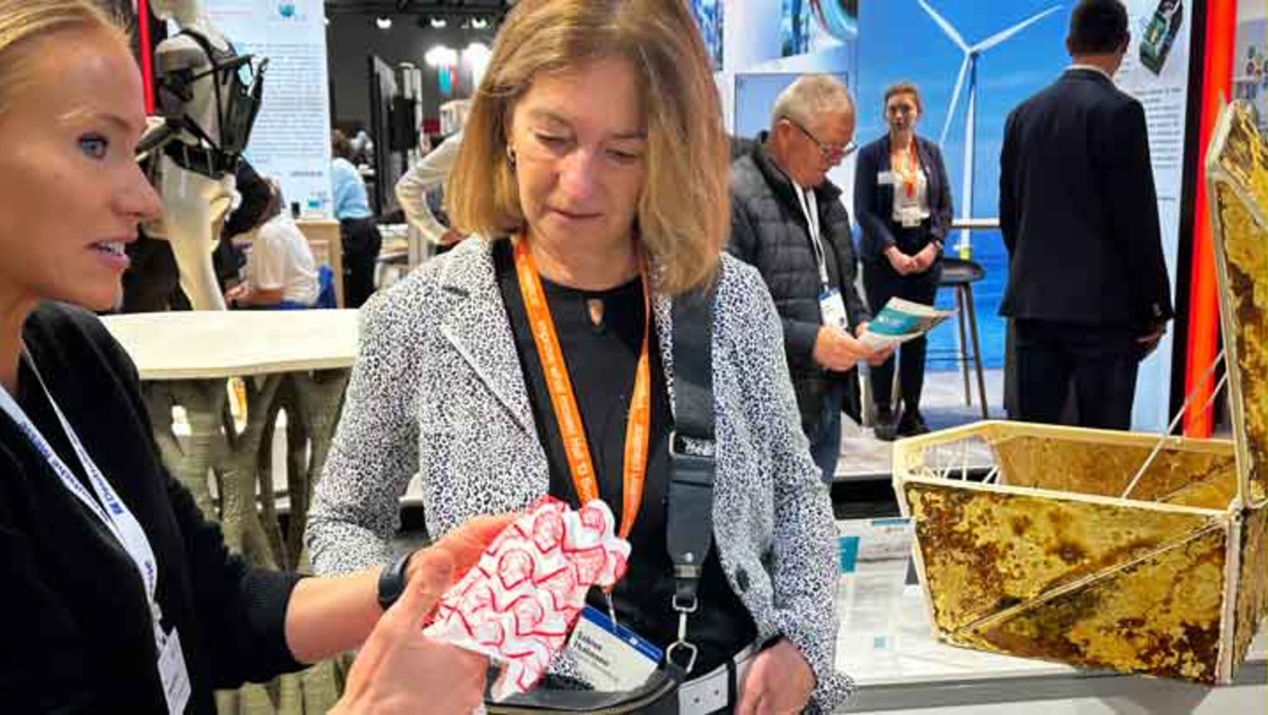 Zwei Frauen auf einer Messe: Eine zeigt der anderen ein rotes, gefaltetes Papiermodell, das wie eine Windkraftanlage geformt ist, während im Hintergrund eine Ausstellung mit Infoständen und Besuchern zu sehen ist.