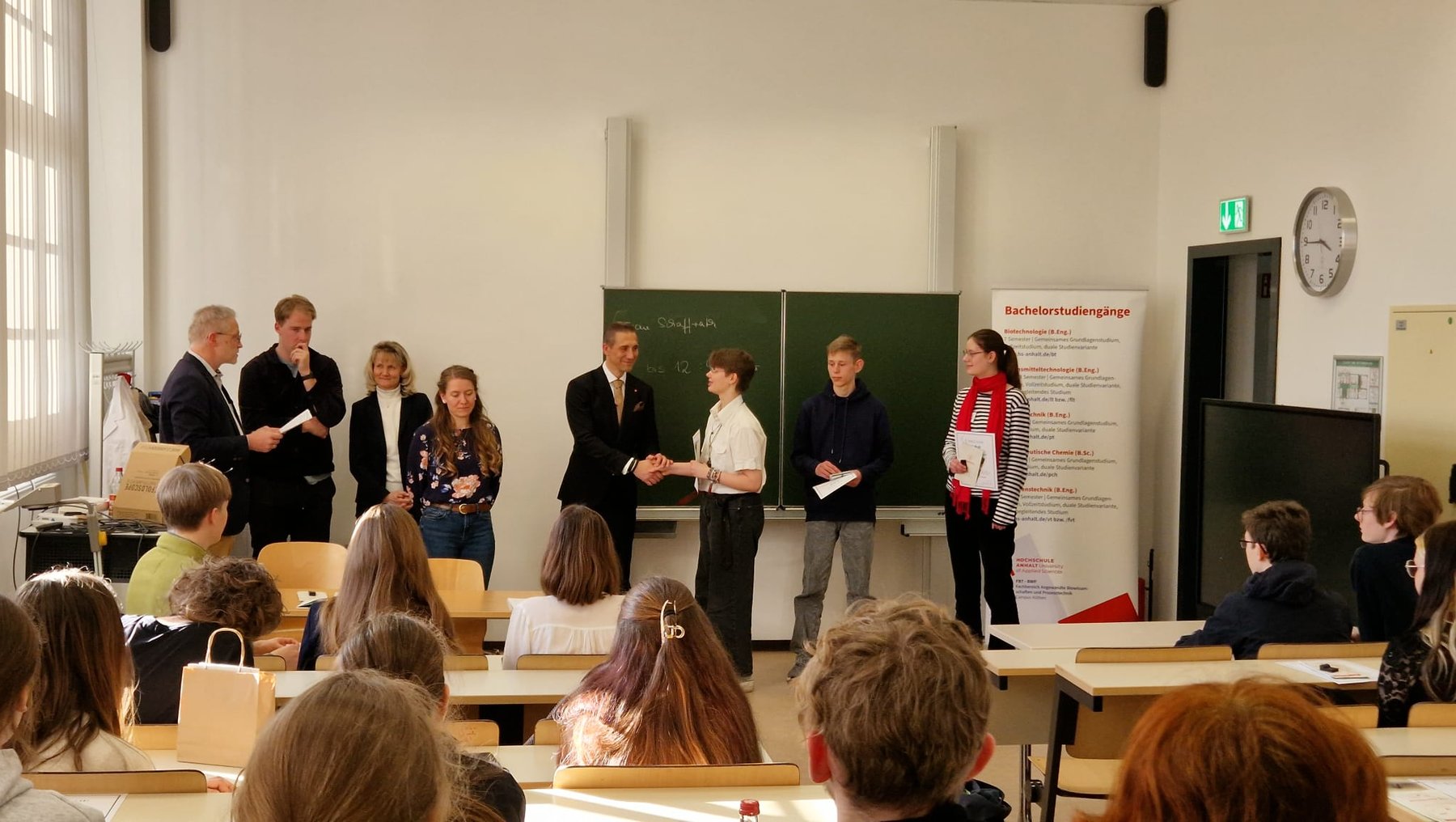 Gruppe von Schülern und Erwachsenen steht im Klassenzimmer vor dem Publikum aus Sitzenden, hält Urkunden oder Preise und wird für eine Leistung ausgezeichnet.