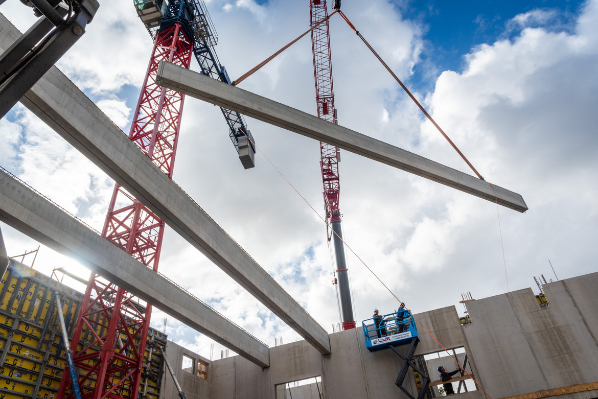 Zwei Arbeiter auf einer Hubarbeitsbühne montieren eine große graue Stahlträgerkonstruktion in einem Rohbau. Im Hintergrund sind zwei rote Baukräne sichtbar, die die Stahlteile halten. Die Baustelle zeigt offene Wände und Deckenöffnungen.