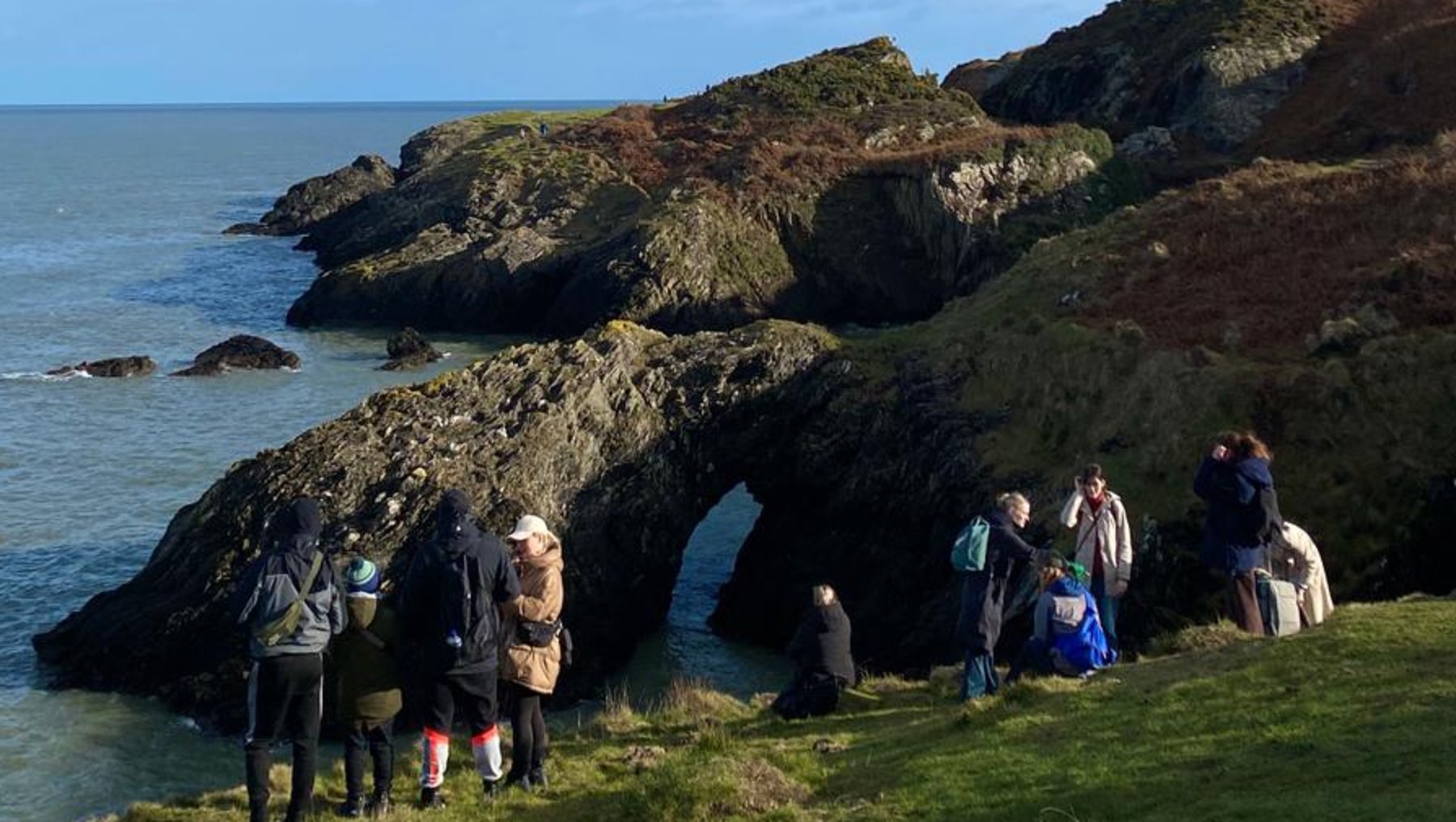 Gruppe von Menschen steht auf einem grasbewachsenen Hang mit Blick auf eine felsige Küste und das blaue Meer bei sonnigem Himmel. Einige Personen beobachten eine Höhlung im Felsen.