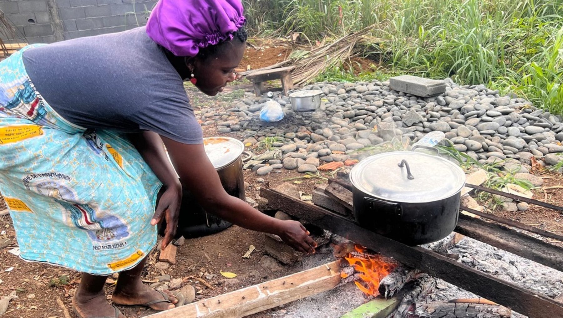 Eine Person in traditioneller Kleidung und lila Kopftuch kocht über einem offenen Feuer mit Holzkohle in einem großen Topf auf einem einfachen Holzgestell.