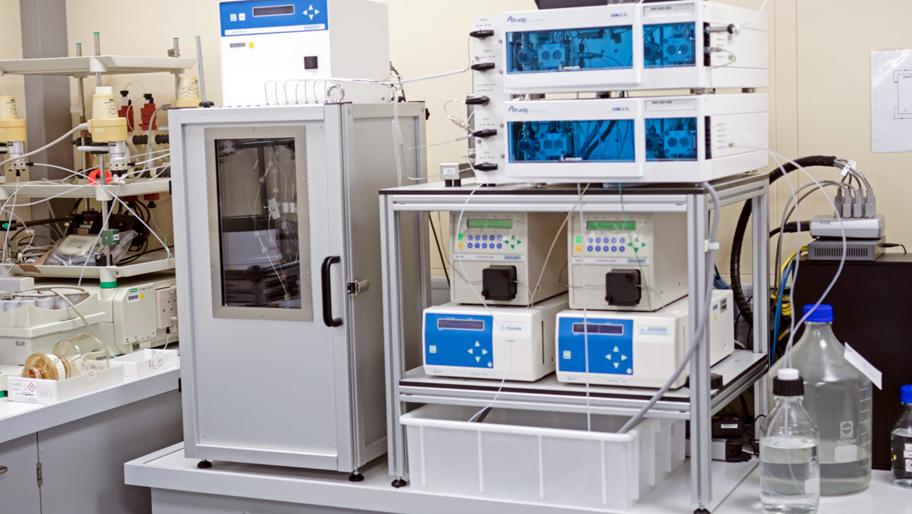 This image shows a lab setup with sophisticated instrumentation. The primary focus includes a fume hood housing equipment with multiple electronic controllers on a frame, likely used for chemical processing or analysis in a laboratory environment. Mechanical arms and containers with liquid are visible.
