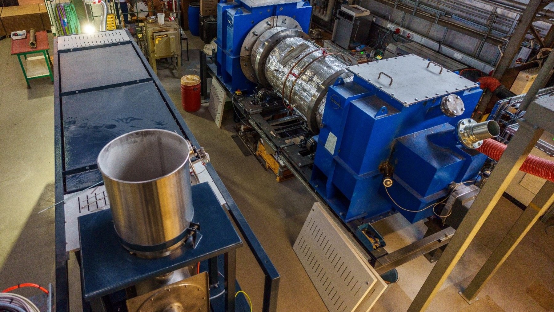 Industrielle Anlage mit großem blauen Turbinenaggregat, das an ein Rohr angeschlossen ist, sowie einem danebenstehenden Edelstahltopf auf einem Heizofen.