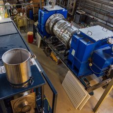 Industrielle Anlage mit großem blauen Turbinenaggregat, das an ein Rohr angeschlossen ist, sowie einem danebenstehenden Edelstahltopf auf einem Heizofen.