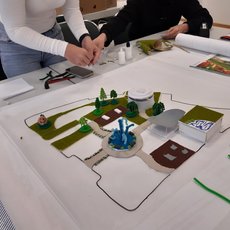 Zwei Personen besprechen gemeinsam ein Modell eines Grundstücks mit Gebäuden, Wegführung und Grünflächen, das auf einem großen Tisch ausgelegt ist.
