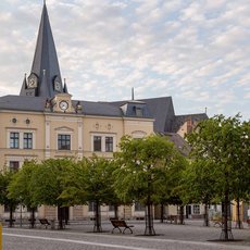 A large, historic building with a clock tower and spire stands prominently in a town square. The building is surrounded by neatly trimmed trees and benches, with a clear sky in the background. The area appears peaceful and well-maintained.