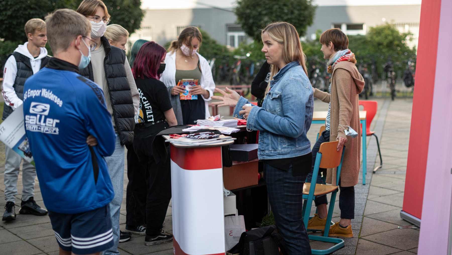 Eine Gruppe von Menschen steht um einen Informationsstand mit rot-weißem Design. Eine Person am Tisch verteilt Flyer oder Prospekte, während andere Interessierte sich bedienen oder warten. Im Hintergrund sind weitere Personen zu sehen.
