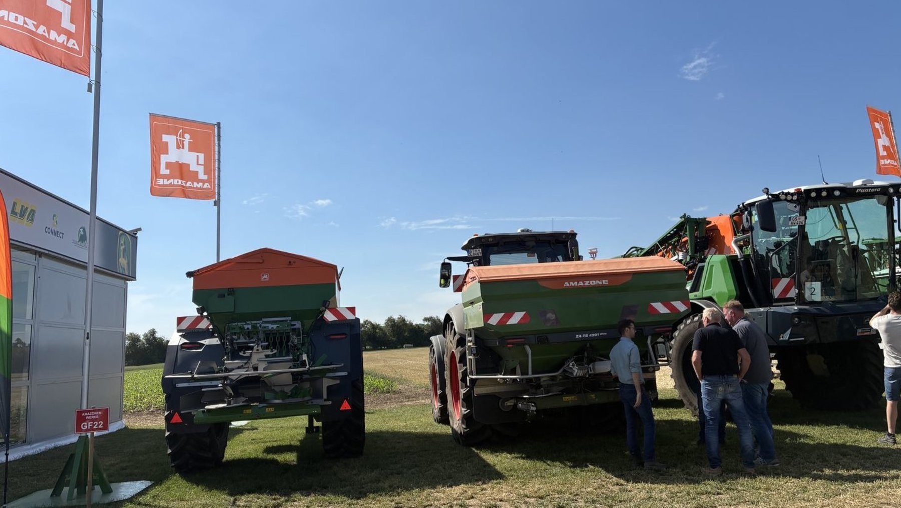 Zwei grüne Landmaschinen, vermutlich Erntemaschinen oder Mähdrescher, stehen auf einem Feld. Drei Personen in Arbeitskleidung stehen in der Nähe und unterhalten sich. Im Hintergrund sind orangefarbene Werbefahnen mit einem Haussymbol zu sehen.