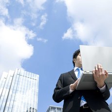 Person im Anzug mit Computer vor Hochhäusern und blauem Himmel