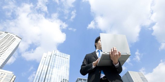 Person im Anzug mit Computer vor Hochhäusern und blauem Himmel