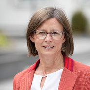 Eine lächelnde Person mit kurz geschnittenen, hellen Haaren und Brille, die ein rotes Sakko über einem weißen Hemd trägt. Im Hintergrund ist eine unscharfe, helle Landschaft mit Gebäuden und Bäumen zu erkennen.