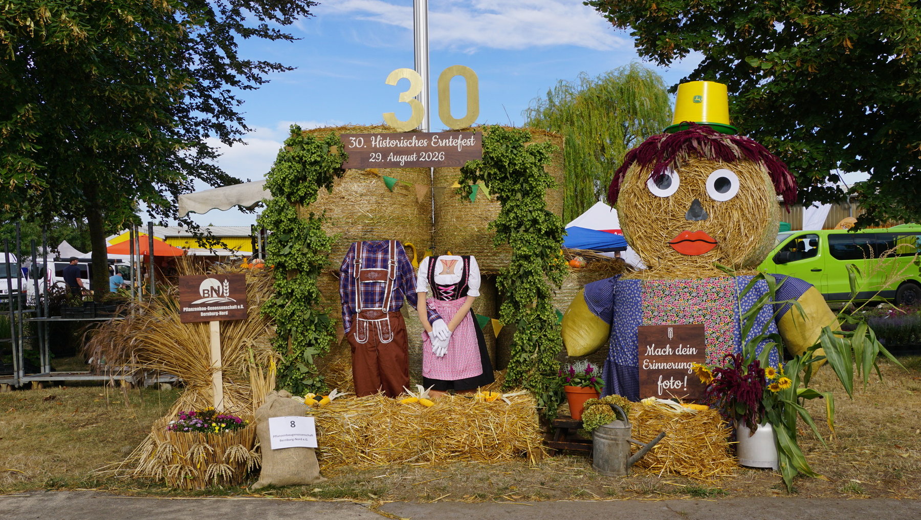 Dekoration zum 30. historischen Erntefest am 29. August 2026 mit Strohfiguren in traditioneller Tracht und einer großen Strohpuppe mit Hut, umgeben von Blumen und Werkzeugen. Im Hintergrund ein Schild mit der Zahl 30 und Festinformationen.
