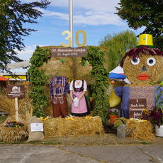 Dekoration zum 30. historischen Erntefest am 29. August 2026 mit Strohfiguren in traditioneller Tracht und einer großen Strohpuppe mit Hut, umgeben von Blumen und Werkzeugen. Im Hintergrund ein Schild mit der Zahl 30 und Festinformationen.
