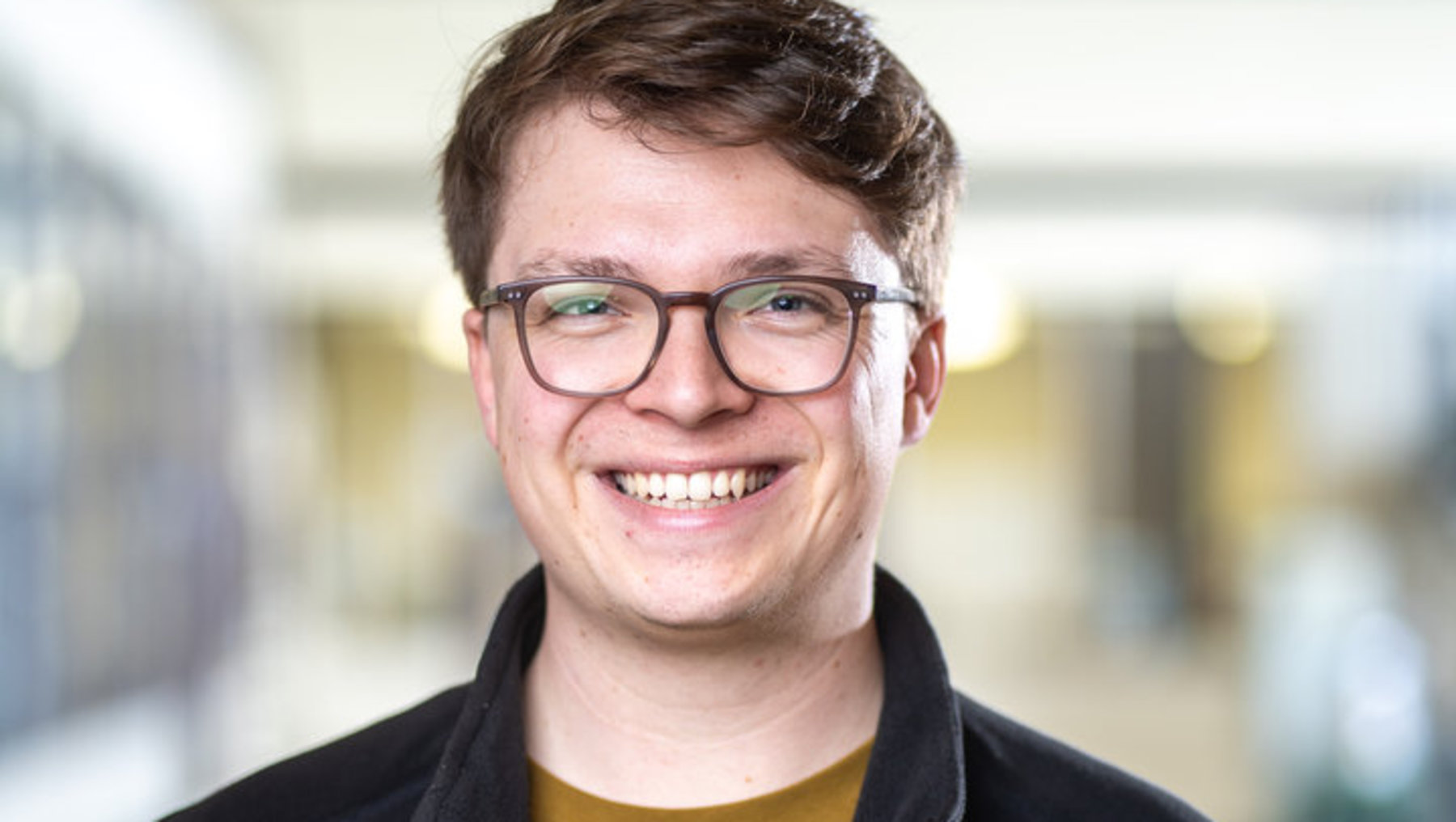 Ein lächelnder Mann mit Brille, kurzem Haar und einem gelben Shirt unter einer schwarzen Jacke steht vor einem hellen Gebäude mit Glasfronten und wirkt freundlich.