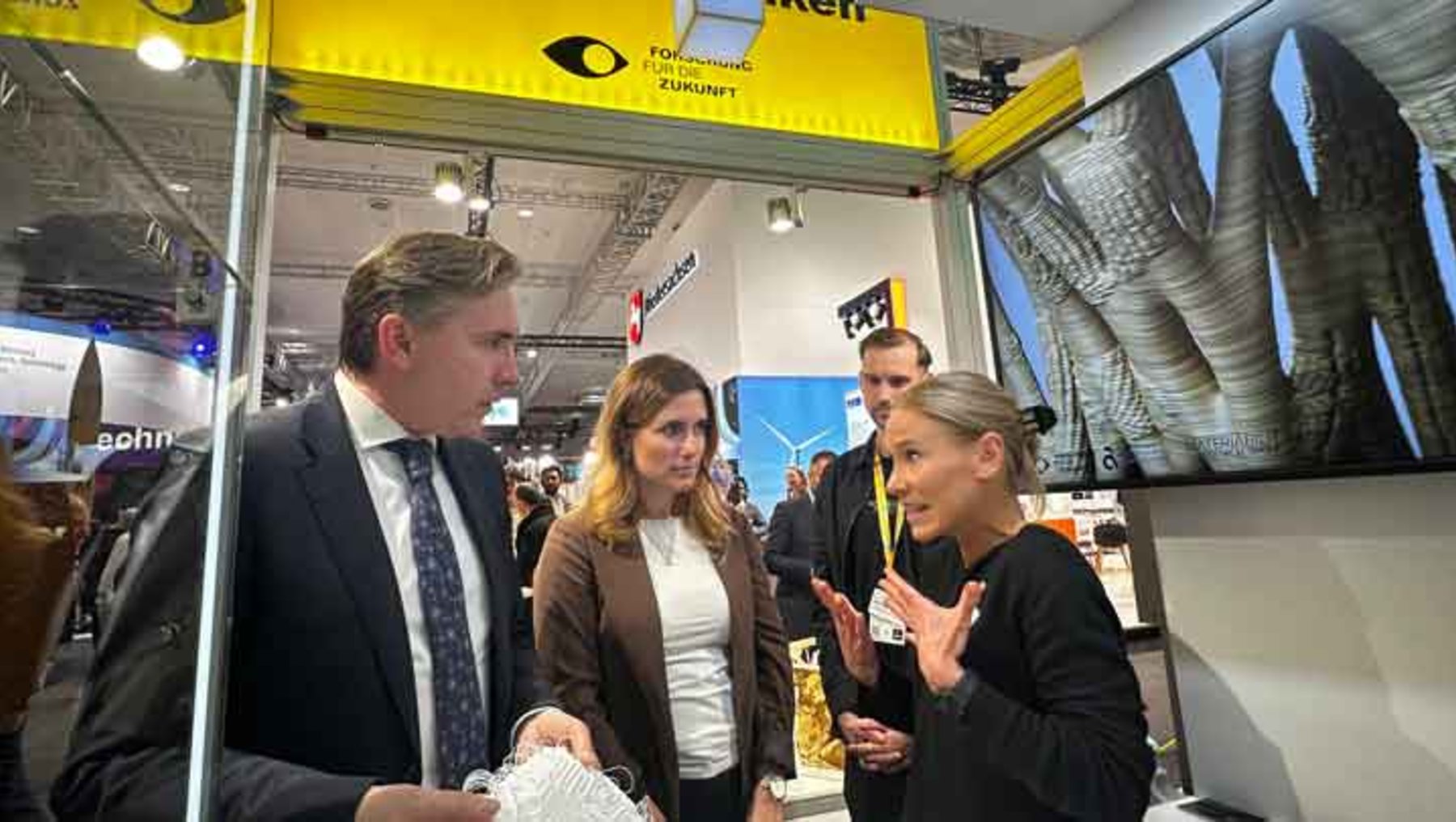 Drei Personen auf einer Messe stehen vor einem Stand mit dem Schriftzug „Fokus Zukunft“. Eine Frau im schwarzen Rollkragenpullover und zwei Männer in Anzug unterhalten sich lebhaft, während im Hintergrund ein großes Bild von Schuhsohlen zu sehen ist.