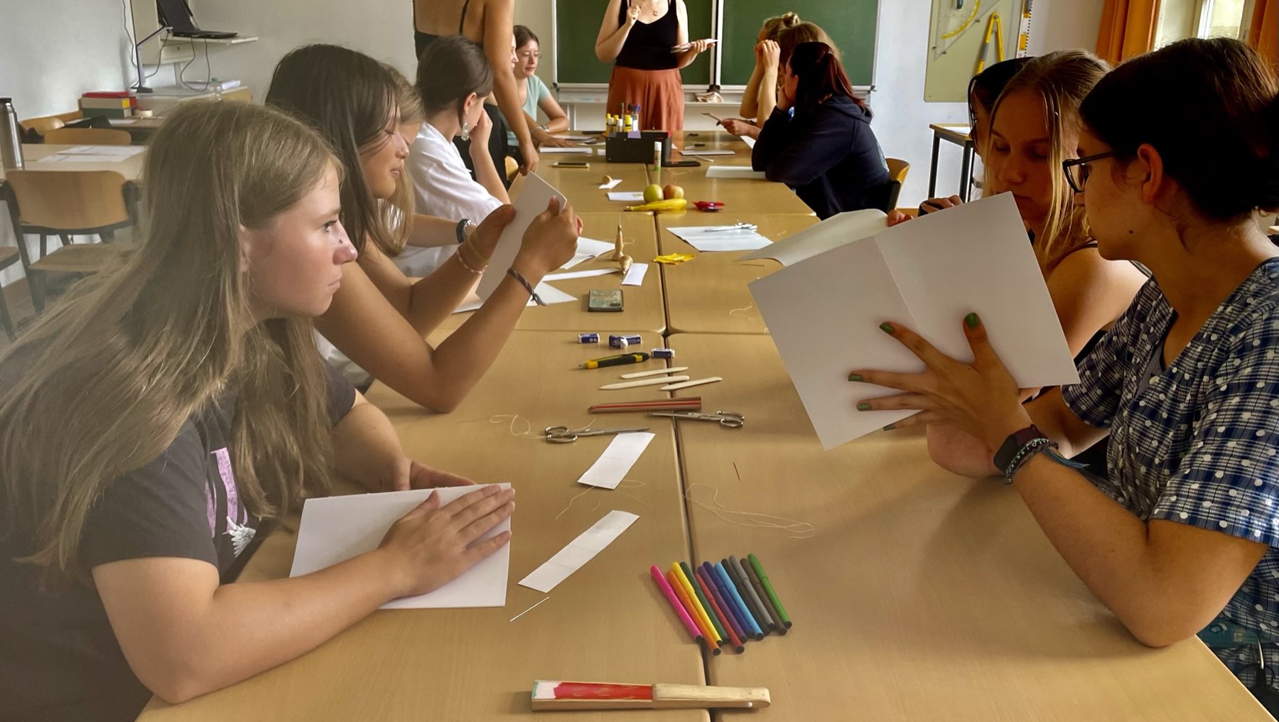 Ein Klassenzimmer mit Schülern und einer Hochschulmitarbeiterin, die gemeinsam an langen Tischen sitzen. Vor ihnen liegen leere Blätter und Stifte, während sie konzentriert arbeiten oder zuhören.