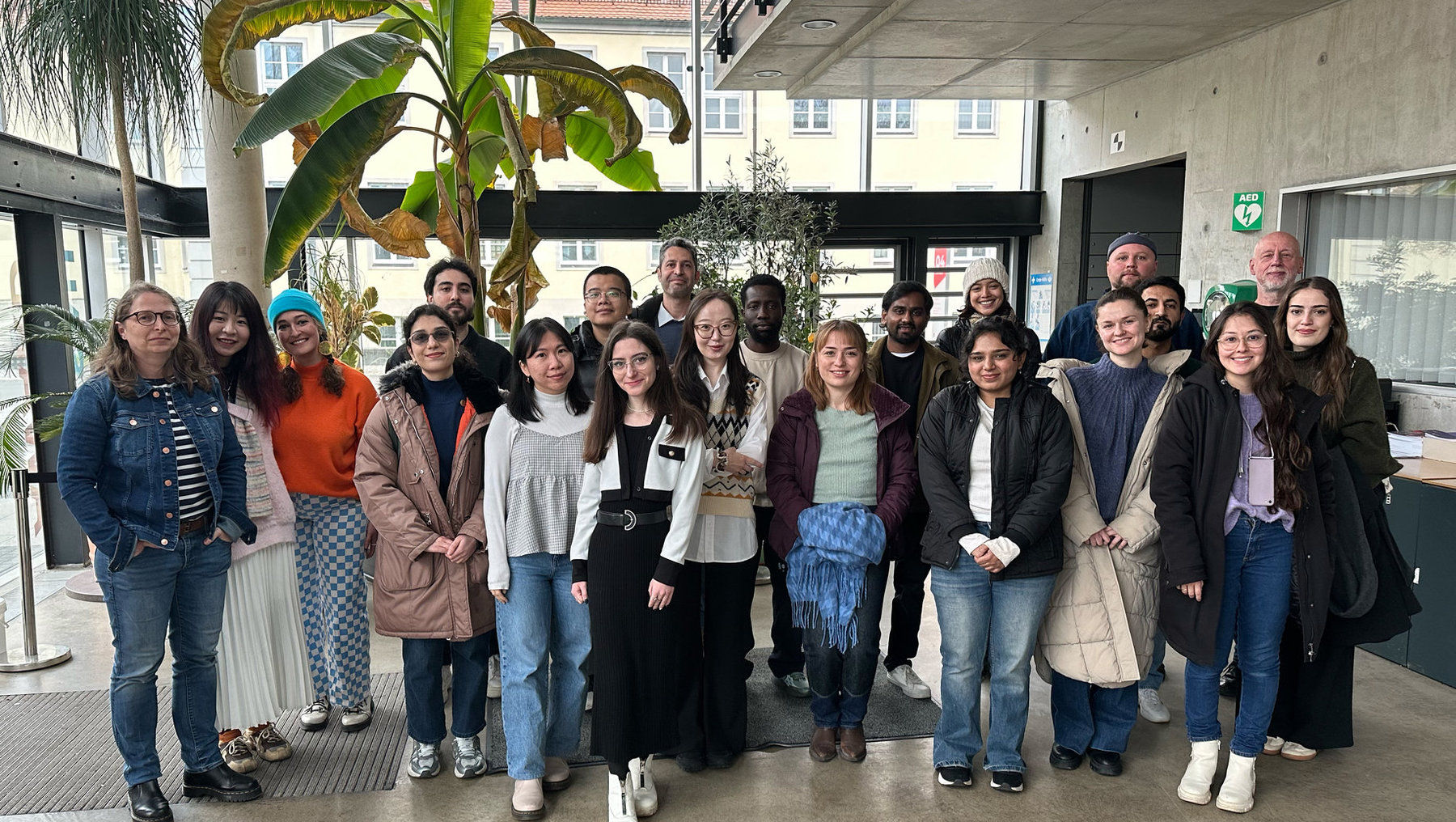 Gruppe von 18 Personen, die vor einem Gebäude mit Pflanze im Hintergrund lächelnd für ein Gruppenfoto posieren. Verschiedene Kleidungsstile, darunter Jacken, Jeans und T-Shirts, sind zu sehen.
